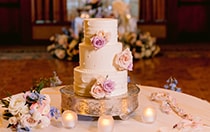 A 3 tiered ornate wedding cake with subtle accents and roses near a bouquet of roses and 3 candles