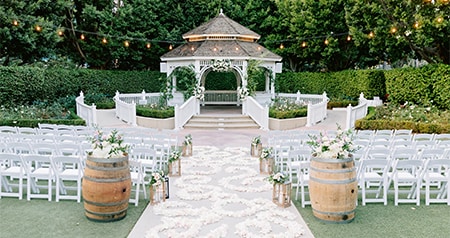 An outdoor ceremony setup with a gazebo, chairs, a flower-lined aisle and string lights overhead	