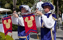 2 performers dressed in 15th century attire play trumpets with Mickey flags attached as a carriage follows behind them