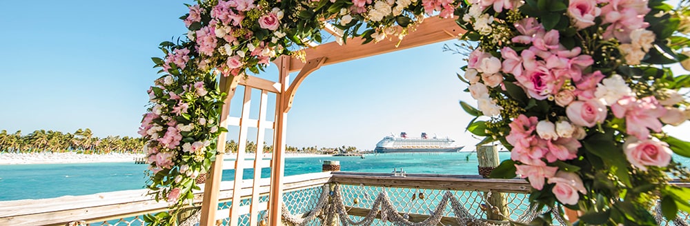 An archway decorated with a floral wreath on a deck in front of the ocean as a cruise ship sails nearby
