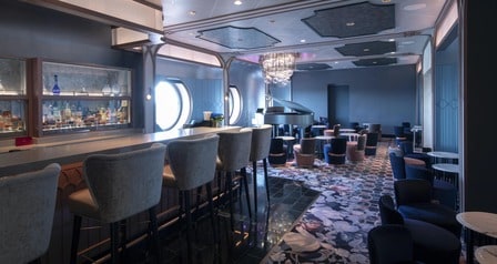 An elegant lounge with bar seating, porthole windows, a crystal chandelier and a grand piano at Nightingale’s aboard the Disney Wish