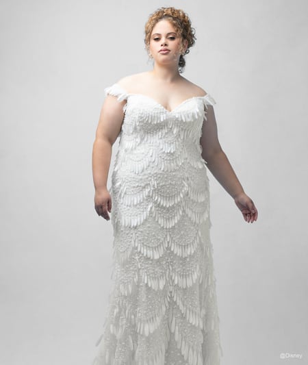 The front of a bride wearing a Tiana inspired sheath gown featuring an off the shoulder neckline and light fringe detail