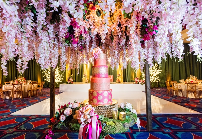 A 4 layer cake sits on a circular table under a canopy of hanging flowers in a room with a dance floor