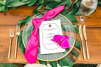 A place setting rests on tropical leaves and consists of 2 forks, 2 knives and a clear plate with an Aulani Resort menu, napkin and flower on top