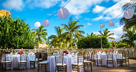 An outdoor reception setup with round tables, chairs and hanging lanterns surrounded by tropical greenery