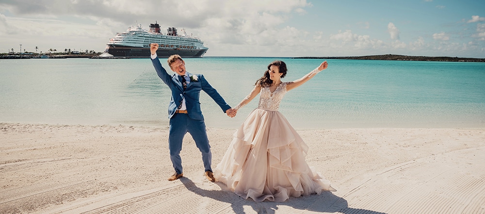 A man in a suit and woman in a wedding dress stand on a sandy beach and hold hands while raising their arms in the air with the ocean and a Disney Cruise Line ship in the background