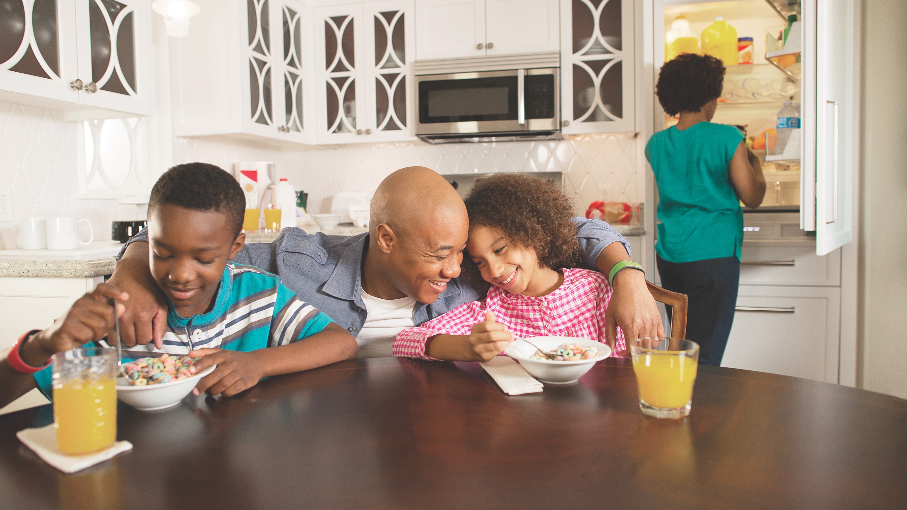 A father hugs his son and daughter while they eat breakfast at a kitchen table