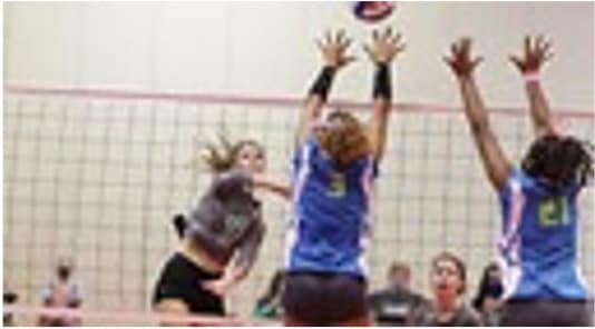 A young volleyball player spiking a volleyball over the net while 2 opposing team players jump with hands raised