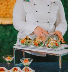 A Disney chef arranging an appetizer table with bowls of salad, Mickey Mouse waffles, fruit skewers, and more snacks in martini glasses