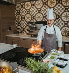 A chef in an apron and hat in an upscale kitchen space sautéing vegetables in a pan with a flame