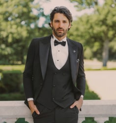 A man in a black tuxedo and bow tie smiling while sitting near floral arrangements  