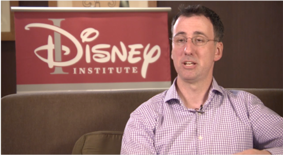 A talking man seated on a couch in front of the Disney Institute sign